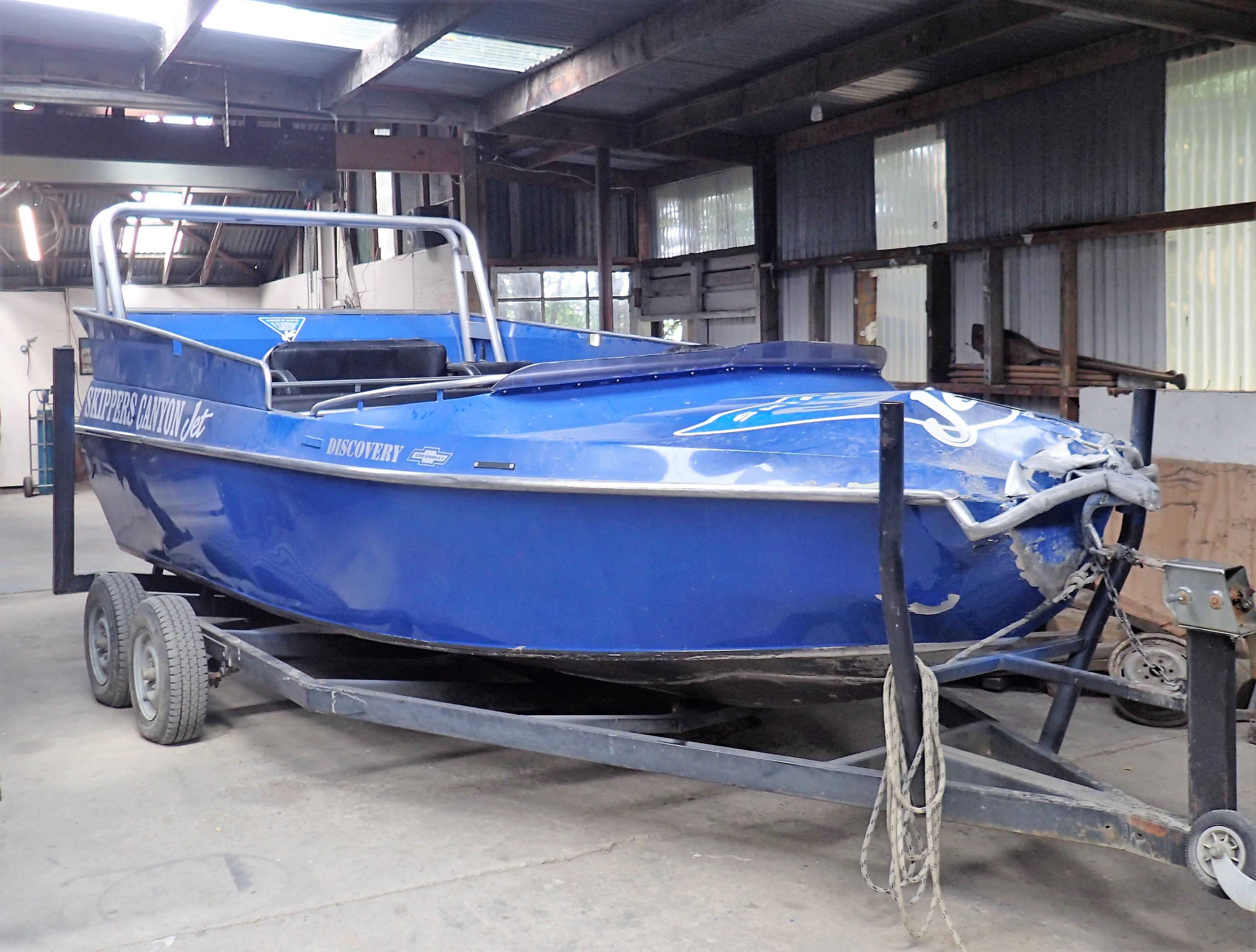 A blue jet boat named Discovery 2 with Skippers Canyon Jet branding sits on a dual-axle trailer inside a shed. The boat has visible damage to its bow, with significant deformation and cracks in the metal structure. The trailer has a winch and rope securing the boat, and the shed has corrugated metal walls and wooden beams. The interior of the boat is visible, showing seating and a roll bar.