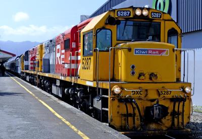 KiwiRail DXC class diesel-electric locomotive number 5287 at a station platform, leading a consist of a second locomotive and passenger carriages. The locomotive is painted in KiwiRail’s yellow, red, grey and white livery, with a large logo on the side.