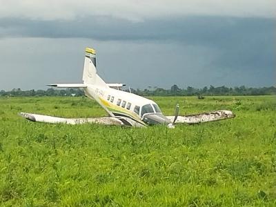 Accident scene showing a PAC-750XL aircraft on a grass field. It has sustained structural damage. Both wings and the fuselage exhibit some deformation and impact damage. The propeller blades are bent rearward, and the main landing gear appears to have collapsed, leaving the aircraft resting on its belly.