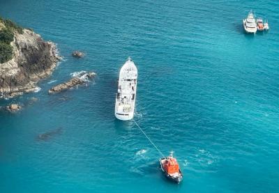 Aerial shot of the Chokyo Maru 68, a 48-metre fishing vessel, after the accident. A line is attached to the stern of the vessel, under tension from a tug. 10-20m off the Chokyo Maru's port beam are prominent coastal rocks both above and below the water surface. Perhaps 80-100m off the Chokyo Maru's starboard bow, three smaller vessels sit in pontoon at anchor.