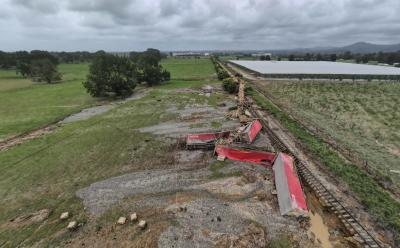 Photo of accident scene. Several container wagons and open laden log wagons lie in disarray in a waterlogged field next to the railway track. Some freight (logs and pulp packaging) is disgorged. The track itself is bent off its embankment. Further wagons stand intact on the track stratching into the distance.