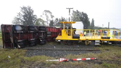 The accident scene. In a light industrial area on a rainy day. The trailer of a large truck and trailer unit lies on its side across railway tracks. There is a large dent in the trailer's perimeter beam. Beyond the trailer, the truck itself stands upright. A brightly painted EU class shunting runner wagon stands upright and derailed. In the foreground is a destroyed 'give way' road sign.