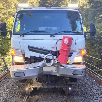 A white Mitsubishi Fuso truck with rail wheels (a Hi-Rail Vehicle) is on a railway track, facing toward the viewer. The front of the truck shows signs of damage, with a deeply crumpled bumper and further damage and dents to the front panels. The truck's headlights are illuminated, and the windshield reflects the sky. The truck is on a track laid with gravel and wooden sleepers, and metal railings are visible on either side of the track. Lush green vegetation forms the backdrop.
