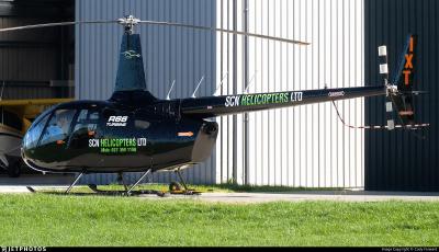 Side view of Robinson R66 Turbine helicopter ZK-IXT parked on a grass apron beside a hangar. The helicopter is painted black with ‘SCN Helicopters Ltd’ markings on the cabin door and tail boom. Main rotor blades are stationary; skids have ground-handling wheels. Tail rotor with black-and-white striped blades is visible, along with ‘IXT’ identification on the vertical fin. Various safety-warning decals are present on the tail boom