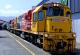 KiwiRail DXC class diesel-electric locomotive number 5287 at a station platform, leading a consist of a second locomotive and passenger carriages. The locomotive is painted in KiwiRail’s yellow, red, grey and white livery, with a large logo on the side.