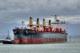The image shows a bulk carrier ship named Thor Nitnirund being guided by a tug traversing The Cut at Port Nelson. The ship is large, almost 200 metres long. The dark hull has visible signs of rust near the waterline, and four prominent yellow gantry cranes mounted on deck. The ship’s superstructure is located toward the stern and bears safety messages painted on it. The sky is overcast, with mountains visible in the background.