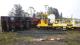 The accident scene. Photo (c) Northern Advocate. The accident scene. In a light industrial area on a rainy day. The trailer of a large truck and trailer unit lies on its side across railway tracks. There is a large dent in the trailer's perimeter beam. Beyond the trailer, the truck itself stands upright. A brightly painted EU class shunting runner wagon stands upright and derailed. In the foreground is a destroyed 'give way' road sign.