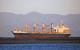  File photo of bulk carrier Achilles Bulker taken in 2022 near Nelson, New Zealand. The vessel is shown high out of the water, with anchor deployed, positioned behind a low-lying gravel or sandbar. Rust staining is visible along the hull, deck cranes, and superstructure. The ship has four cargo cranes and closed hatch covers, indicating a geared bulk carrier configuration. The vessel is lit by sunlight from a low angle, casting warm tones across the scene. Mountain ranges are visible in the background.