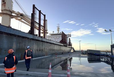 Two accident investigators wearing high-visibility gear and safety helmets approach a large bulk carrier berthed at a port. The vessel's hull shows signs of rust, and the front of the main superstructure bears the stencilled words "SAFETY FIRST." In the foreground, several hose handling trolleys are lined up on the wharf. The concrete surface of the wharf is puddled and reflective after rain, with orange and white safety cones placed nearby. The sky is mostly clear and the Sun is low, behind the bulk of the