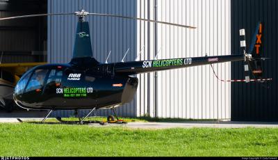 Side view of Robinson R66 Turbine helicopter ZK-IXT parked on a grass apron beside a hangar. The helicopter is painted black with ‘SCN Helicopters Ltd’ markings on the cabin door and tail boom. Main rotor blades are stationary; skids have ground-handling wheels. Tail rotor with black-and-white striped blades is visible, along with ‘IXT’ identification on the vertical fin. Various safety-warning decals are present on the tail boom