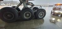 Fig 4 from the report shows the damaged right landing gear with deflated tyre. Photo Air New Zealand Close-up view of the right-hand main landing gear of a Boeing 777 aircraft on a wet airport apron during rainfall. The image shows six main gear tyres; the rear-most tyre (right side of image) is visibly deflated and compressed under load, with sidewall deformation. Other tyres appear fully inflated. Water runoff is visible on the gear structure and pooling on the concrete surface. A ground vehicle is present in the background to the right.