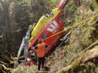 A TAIC investigator surveys the scene. Photo NZDF The wreckage of a crashed BK117 rescue helicopter sits upright in dense bush on a steep slope. The helicopter has a red fuselage with 'RESCUE' and 'King Country – Coromandel' markings, and a yellow tail boom. the main rotor blades are torn up, ripped and distorted from slashing through the trees during the crash sequence. A man in a high-visibility vest bearing the TAIC logo stands with his back to the viewer, looking at the wreck. Another person in rescue gear is partially visible among the vegetation.