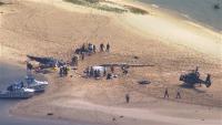 Aerial photo of the accident scene. On a sandy beach, -- a sandy beach where one EC130 helicopter sit upright and apparently intact. A few dozen metres away and just above the calm waterline, the fuselage of another EC130 lies on its side, partly embedded in the sand. Between the two helicopters lies the main rotor head and rotor blades of the wrecked helicopter. Around two dozen people including emergency first responders are present. 