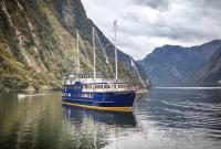 A large blue and yellow passenger vessel named 'Fiordland Navigator' is anchored on calm water in a mountain fjord. The vessel has three tall masts, each with a furled jib sail for optional wind-powered sailing. It features three decks with rows of windows, and an open front viewing platform. In the background, a small craft moves across the water. The fjord is surrounded by steep, forested mountains under a cloudy sky.