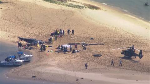 Aerial photo of the accident scene. On a sandy beach, -- a sandy beach where one EC130 helicopter sit upright and apparently intact. A few dozen metres away and just above the calm waterline, the fuselage of another EC130 lies on its side, partly embedded in the sand. Between the two helicopters lies the main rotor head and rotor blades of the wrecked helicopter. Around two dozen people including emergency first responders are present. 