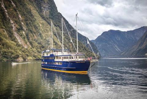 A large blue and yellow passenger vessel named 'Fiordland Navigator' is anchored on calm water in a mountain fjord. The vessel has three tall masts, each with a furled jib sail for optional wind-powered sailing. It features three decks with rows of windows, and an open front viewing platform. In the background, a small craft moves across the water. The fjord is surrounded by steep, forested mountains under a cloudy sky.