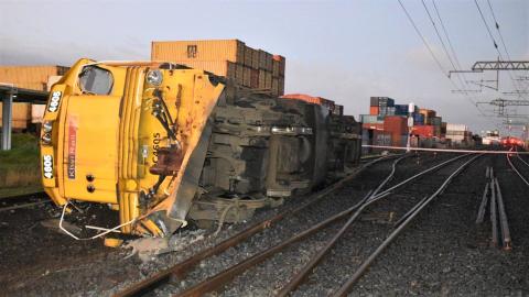 A rail locomotive lies on its side beside a set of railway points. Railway power lines are overhead, intact. In the distance are shipping containers stacked in land adjoining the rail corridor. 
