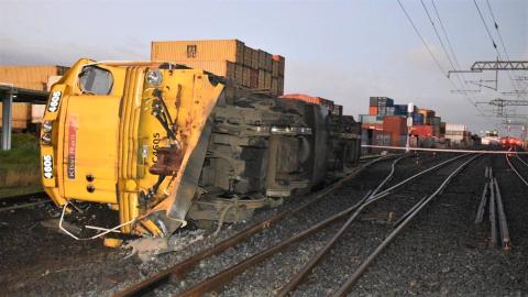 The locomotive, DC4605 lies on its side at the accident site | Photo credit Darren Masters via NZ Herald A rail locomotive lies on its side beside a set of railway points. Railway power lines are overhead, intact. In the distance are shipping containers stacked in land adjoining the rail corridor.